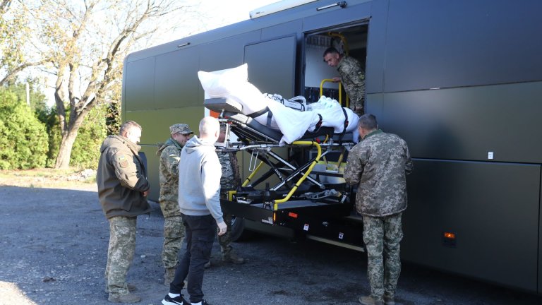 Fire menn i militær uniform og en mann i sivil løfter en båre inn i buss