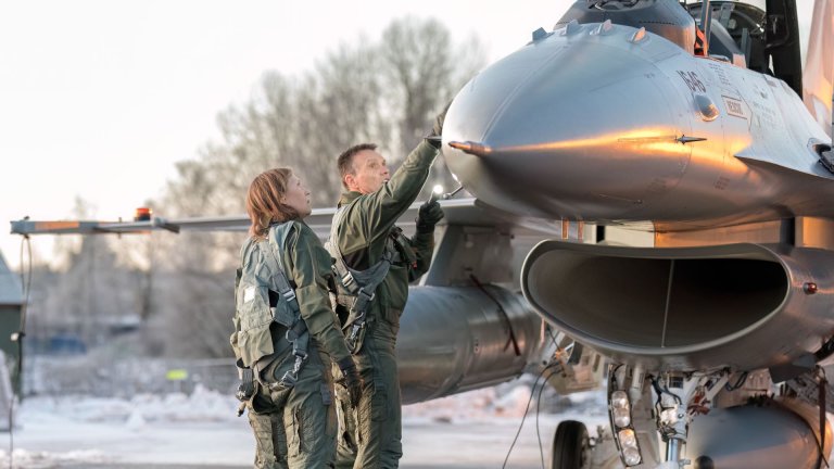 En mann og en kvinne i flygeruniform inspiserer et jagerfly med vinterkledd landskap i bakgrunnen.