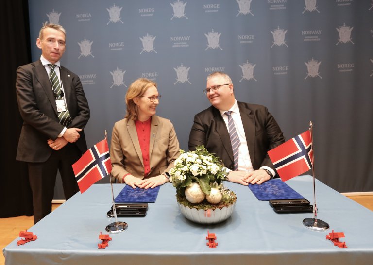 Tre personer i formelle klær deltar i en signeringsseremoni foran en blå bakgrunn med Norges Forsvars emblem. To personer sitter ved et bord med norske flagg og en blomsterdekorasjon, mens den tredje står ved siden av og ser på.