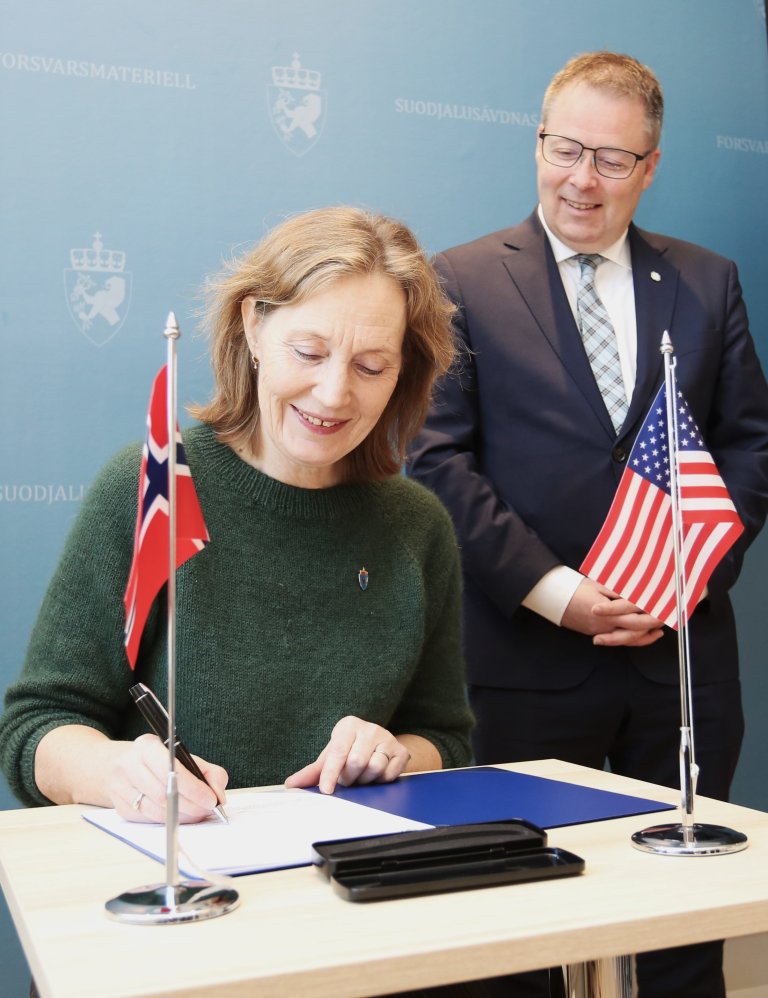 Smilende kvinne i grønn genser signerer papirer ved et bord med et norsk og et amerikansk flagg, mens smilende mann ikledd dress og slips står smilende bak.