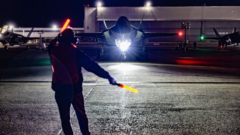 En handler med lyspinner står med ryggen mot kamera og veileder et F-16 fly som står ved en hangar på en flyplass.