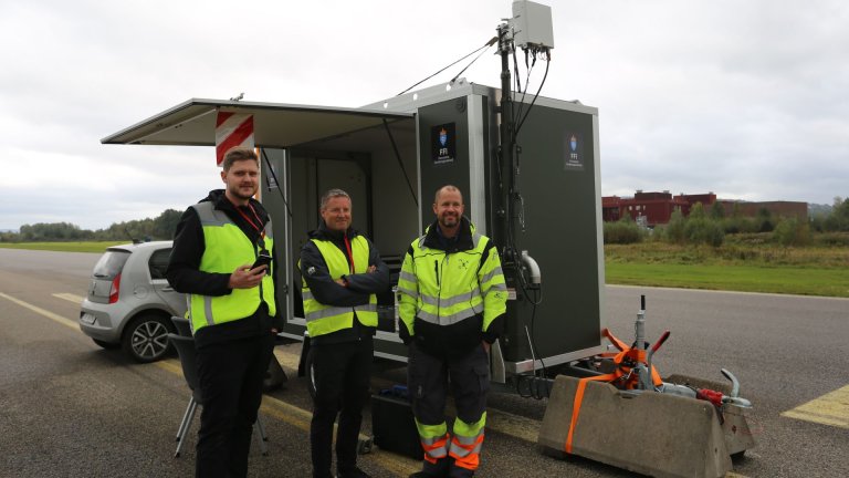 Tre menn foran en skaptilhenger med utstyr på flystripe