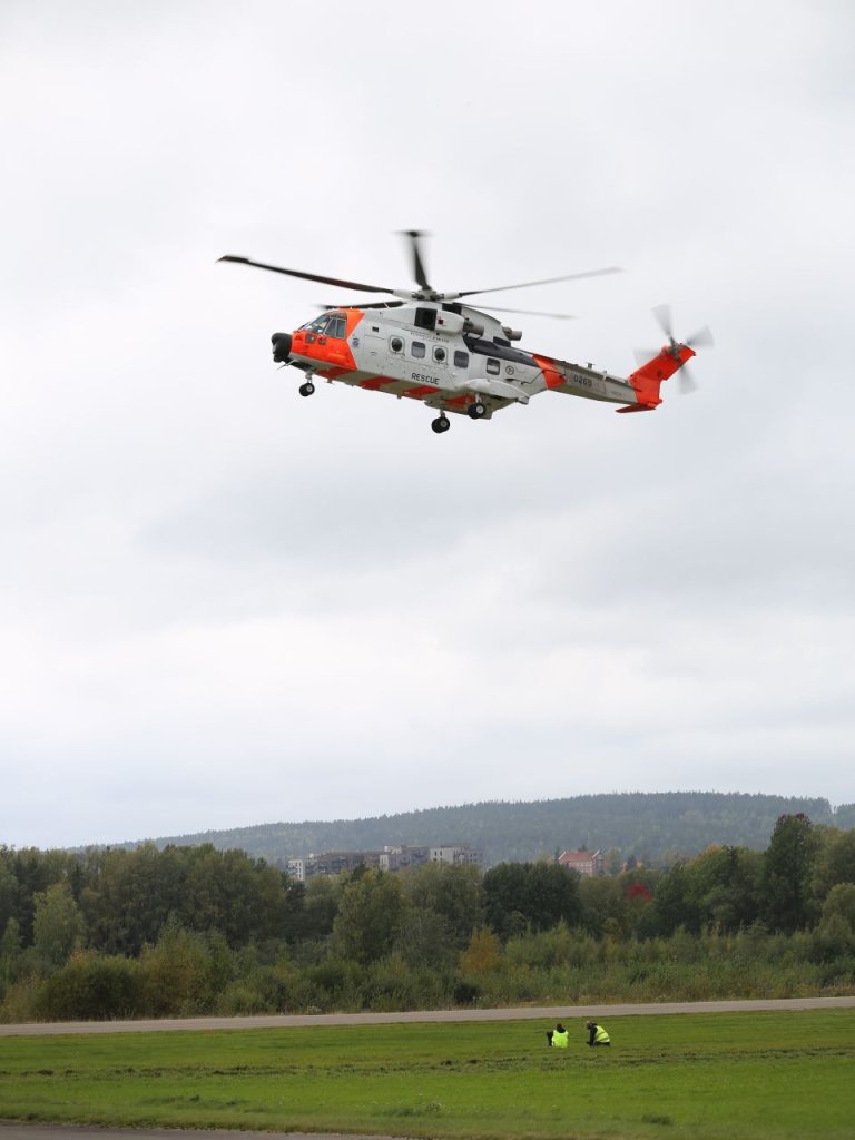 Redningshelikopter i luften over to personer i gul vest som sitter på bakken