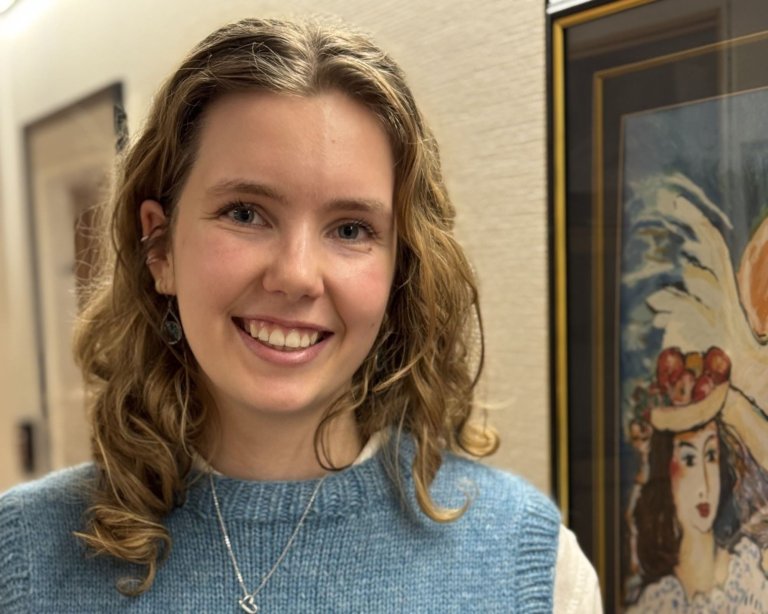 En ung kvinne med krøllete, lysebrunt hår smiler mot kameraet. Hun har på seg en lyseblå ullvest over en hvit topp og står innendørs ved siden av et innrammet maleri som viser et portrett av en kvinne med frukter og fjær på hodet.