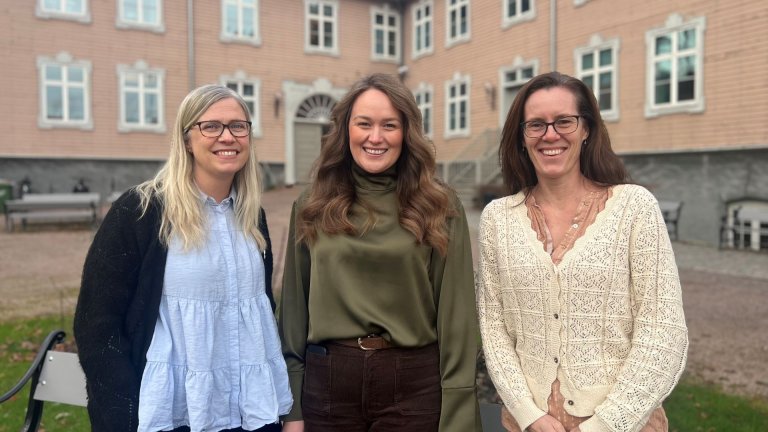 Tre kvinner smiler foran en lys trebygning