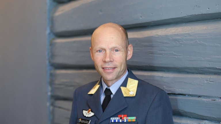 En mann i norsk luftmilitær uniform står foran en blågrå tømmervegg og ser rett mot kameraet med et smil. Uniformen har distinksjoner på kragen og flere dekorasjoner på brystet.