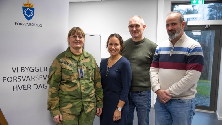 To kvinner, hvorav en i militæruniform og to mann står smilende mot fotografen foran en banner med Forsvarsbyggs logo.