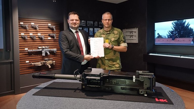 To menn holder en signert kontrakt foran et tungt våpensystem i et utstillingsrom med skytevåpen på veggen.