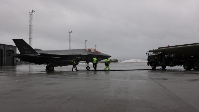 Et kampfly av typen F-35 står parkert på en våt flyplass med grått og overskyet vær. Tro personer i refleksvester står ved flyet med en slange og fyller drivstoff fra en tankbil