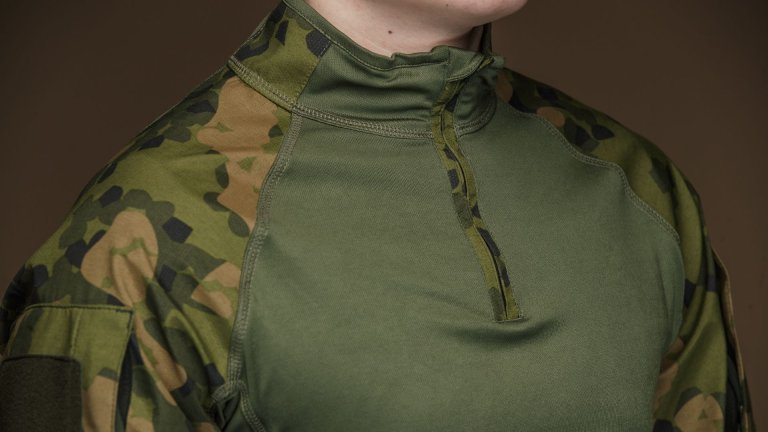 Et nærbilde av overkroppen til en person iført en militær trøye med grønt stoff og kamuflasjemønster på skuldrene og ermene.