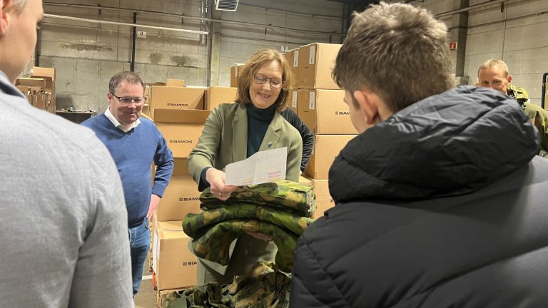 Smilende og pent kledd kvinne gur en bunke med uniformstøy til en ung mann, ved siden av henne står en smilende mann. De står i en lagerhall med mange pappesker stablet opp i bakgrunnen.