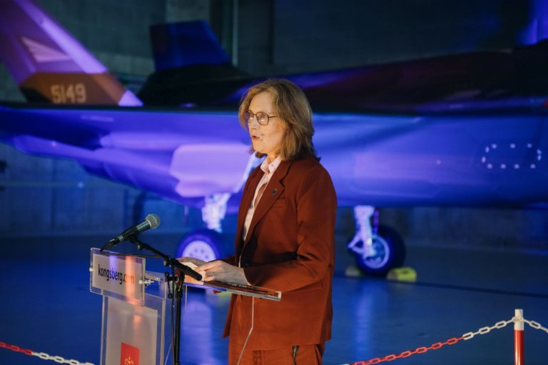 Kvinne snakker ved en talerstol med et kampfly i bakgrunnen.