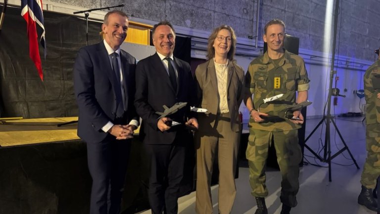 To menn i dress, en kvinne i dress og en soldat i uniform står smilende foran en scene, mens to av dem holder modeller av kampfly og missiler