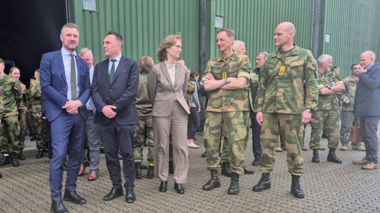 To sivile menn i dress, en kvinne i beige dress og to soldater i uniform står samlet i samtale utenfor en grønn lagerbygning, med flere mennesker i militæruniform i bakgrunnen
