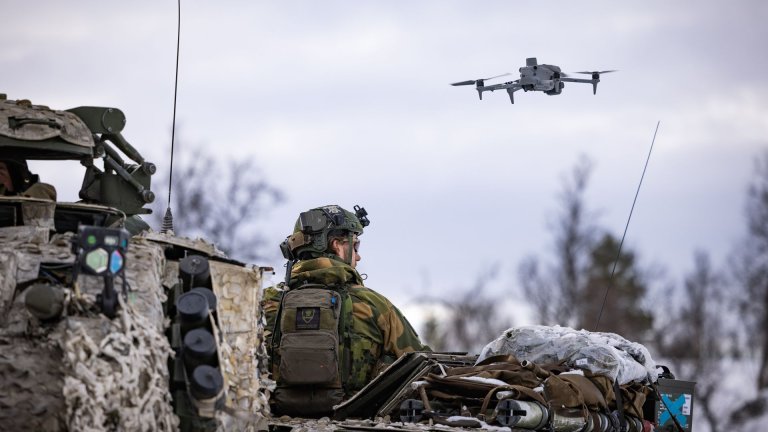 Soldat i kamuflasje sitter på et militært kjøretøy mens en drone flyr over.