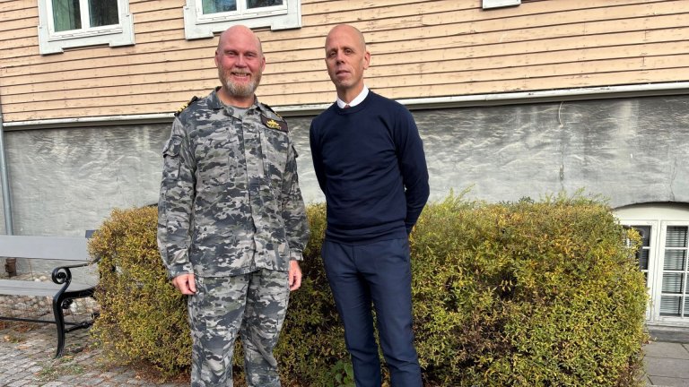 To menn står foran en bygning. Den ene er iført marineuniform med kamuflasjemønster, den andre er kledd i mørk genser og dressbukse.