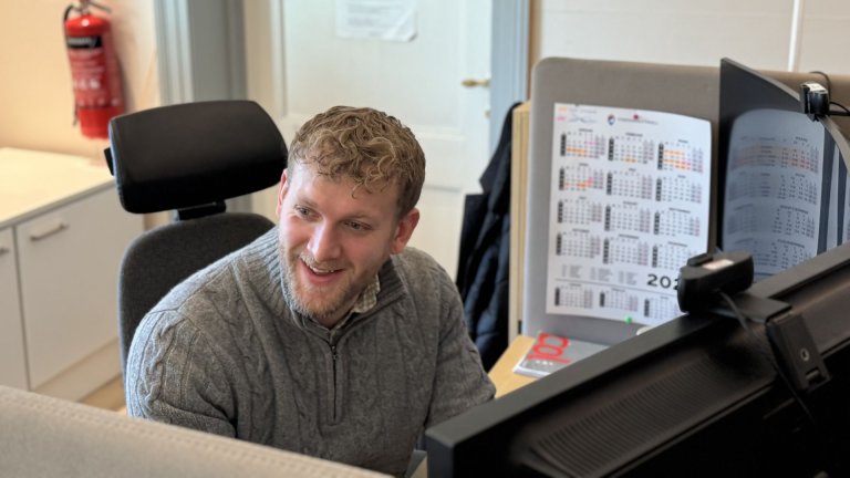 Smilende mann sitter ved en kontorpult foran dataskjermer